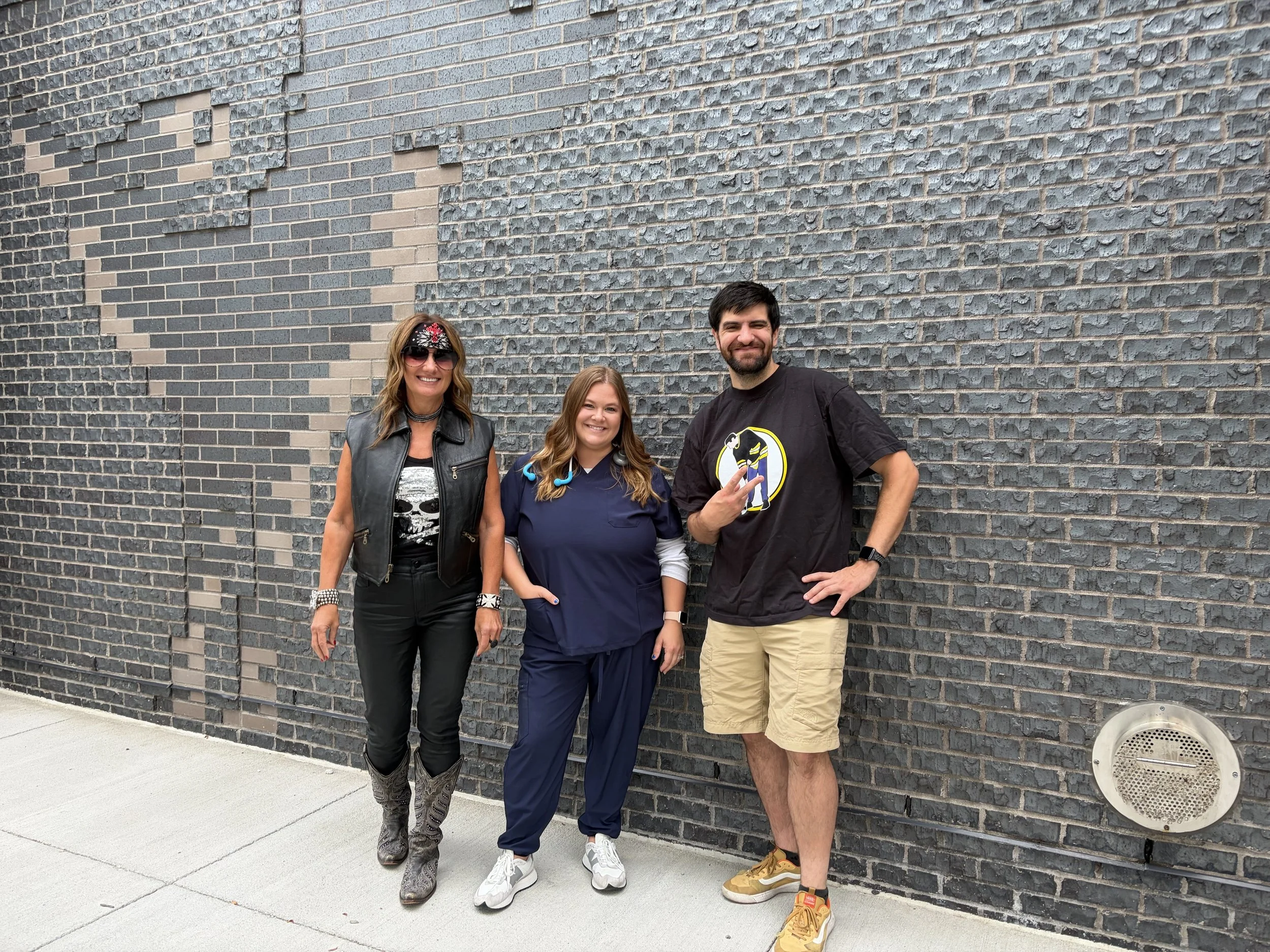 Three people standing in front of a brick wall. The woman on the left is wearing sunglasses, a bandana, a leather vest, and black pants. The young girl in the middle is wearing blue scrubs with headphones around her neck. The man on the right is wear