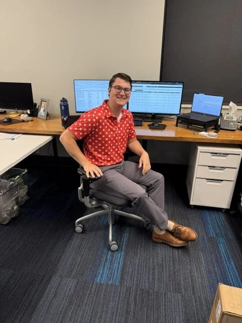 Young man sitting at office desk with two computer monitors, a laptop, and office supplies, smiling at the camera.