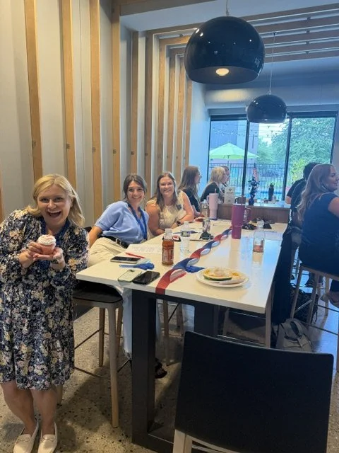 Group of women sitting at a table in a restaurant, with one girl holding a cupcake and smiling at the camera, others chatting and dining.