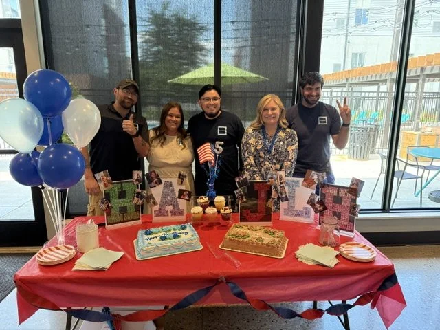Five people standing behind a table with birthday decorations, cakes, and balloons, celebrating indoors with large windows showing an outdoor patio area.