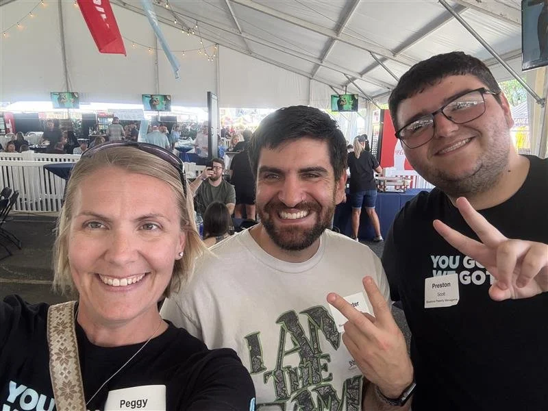 Three people smiling for a selfie inside a large tent at an event, with hanging decorations and screens in the background. The woman on the left is named Peggy, and the man on the right is named Preston.