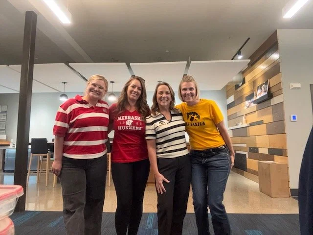 Four women standing together in an indoor space, smiling at the camera. Three of the women are wearing Nebraska Huskers shirts, and one is wearing a red and white striped top.