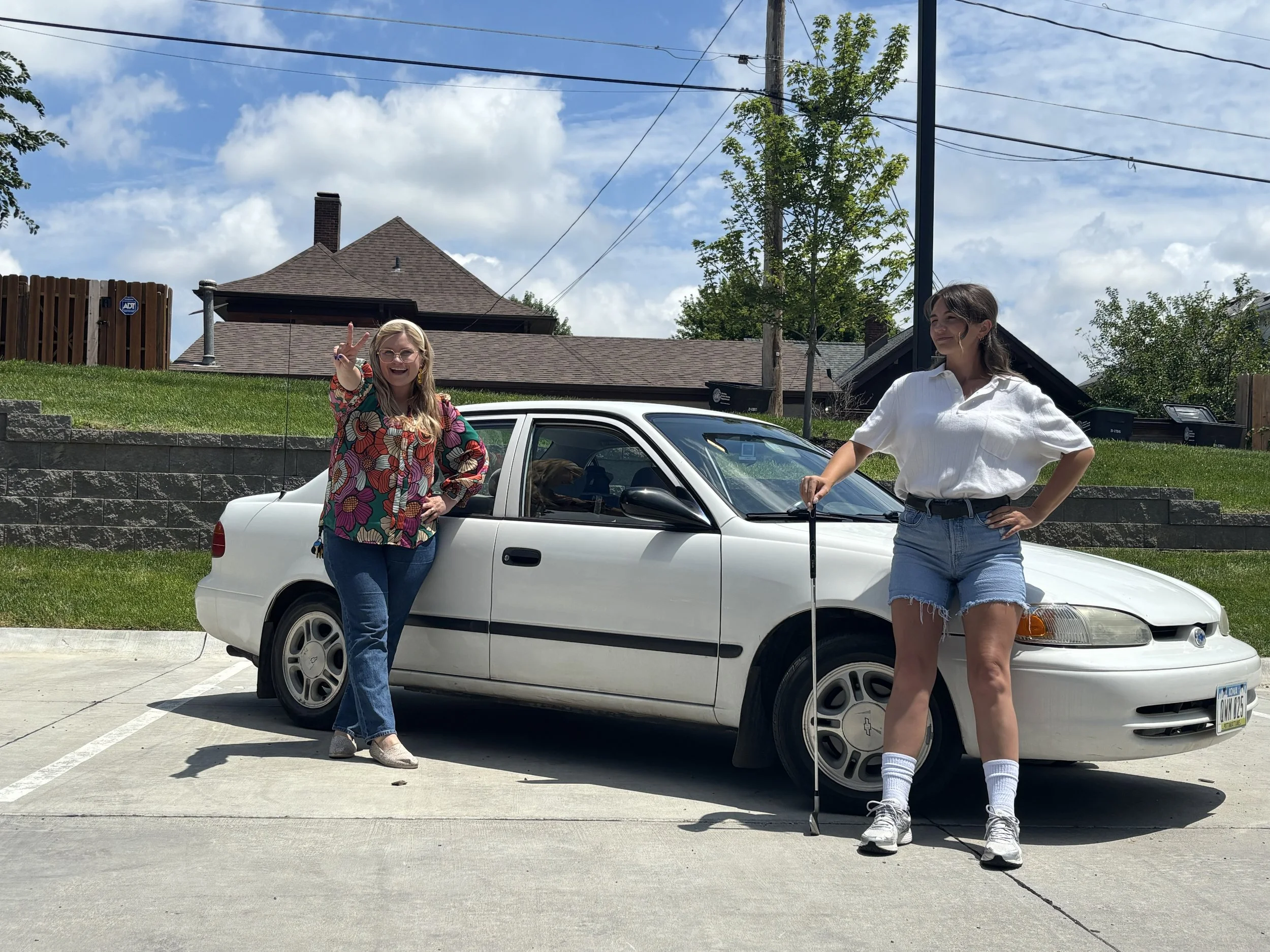 Two women standing beside a white car parked in a driveway on a sunny day. One woman with blonde hair, wearing glasses and a colorful floral top, is smiling and making a peace sign. The other woman with dark hair, wearing a white shirt and denim shor