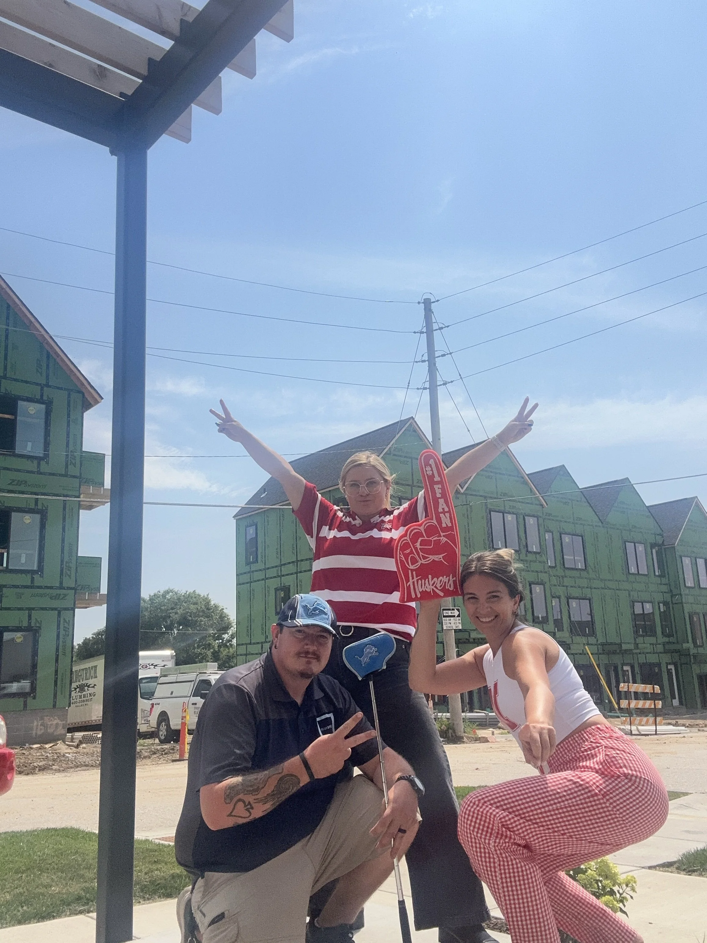 Three young adults posing outdoors under a clear blue sky, with house construction in the background. The woman in the red and white striped shirt is making a peace sign, the man with tattoos is crouching and holding a tiny flag, and the woman in che