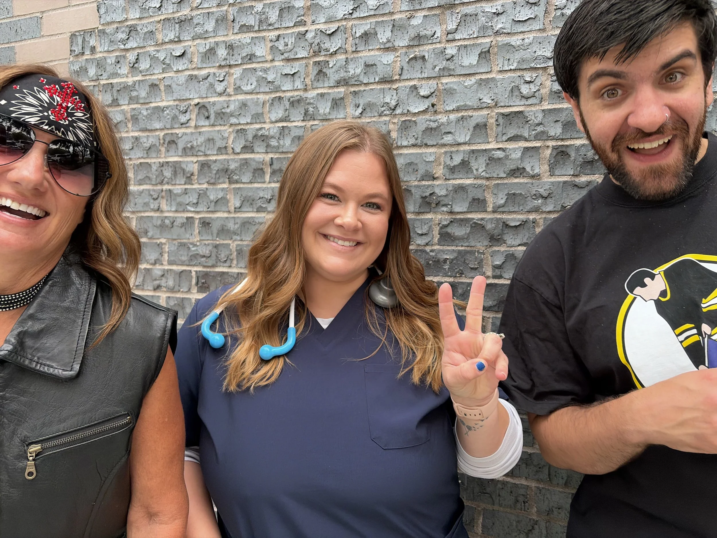 Close-up of three people smiling and making peace signs in front of a gray brick wall. The left wears sunglasses and a black bandana with red embroidery, the middle has long brown hair and wears medical scrubs with a stethoscope, and the right has da