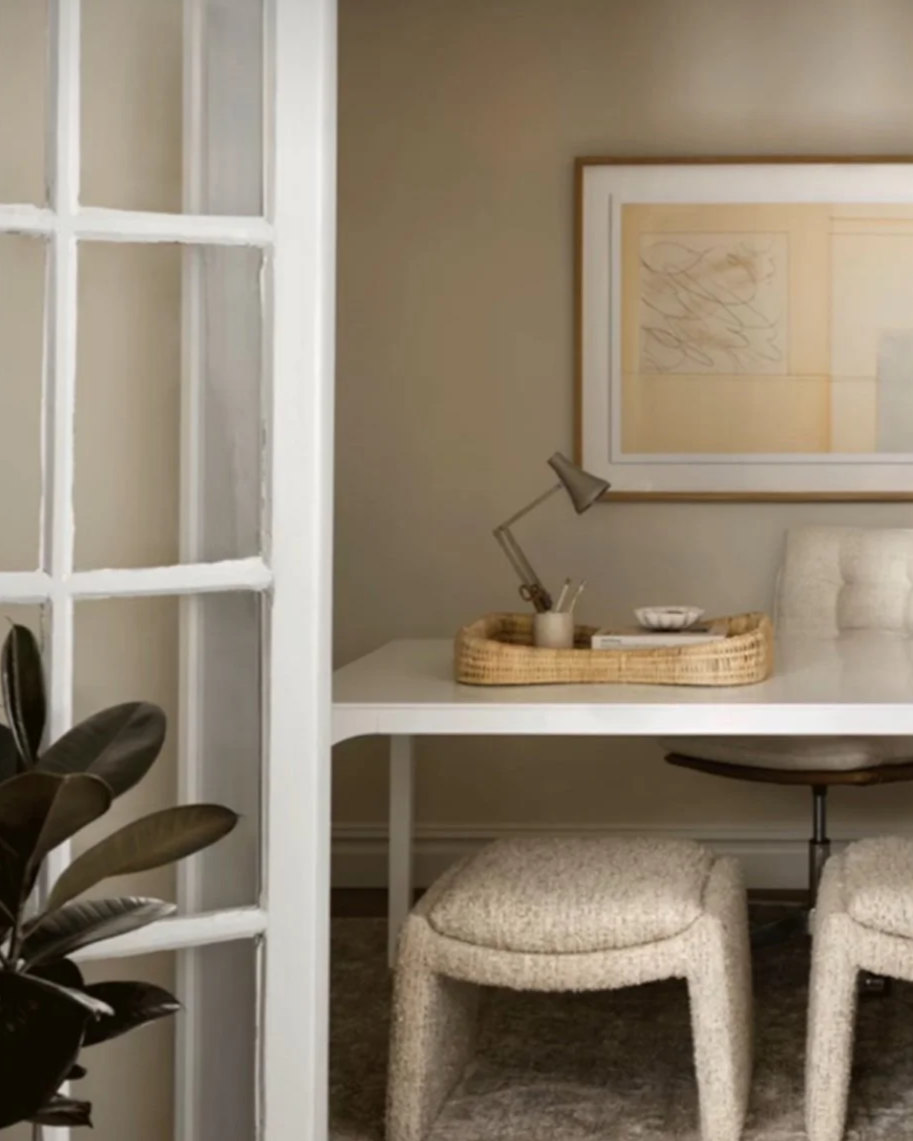 A cozy workspace with a white desk, beige upholstered stools, a framed abstract art piece on the wall, a small desk lamp, and a wicker tray holding stationery, seen through a glass-paneled door.
