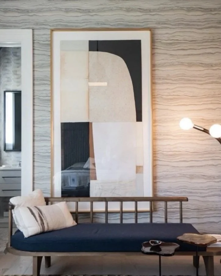 Interior of a modern living room with a wooden sofa, decorative pillows, a framed abstract art piece on textured wall, and a side table with books and a remote.