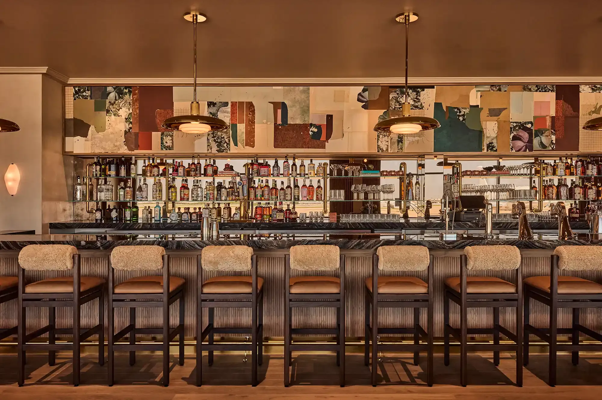 A bar counter with high chairs, shelves of various liquor bottles, and decorative wall art in the background, illuminated by pendant lights.