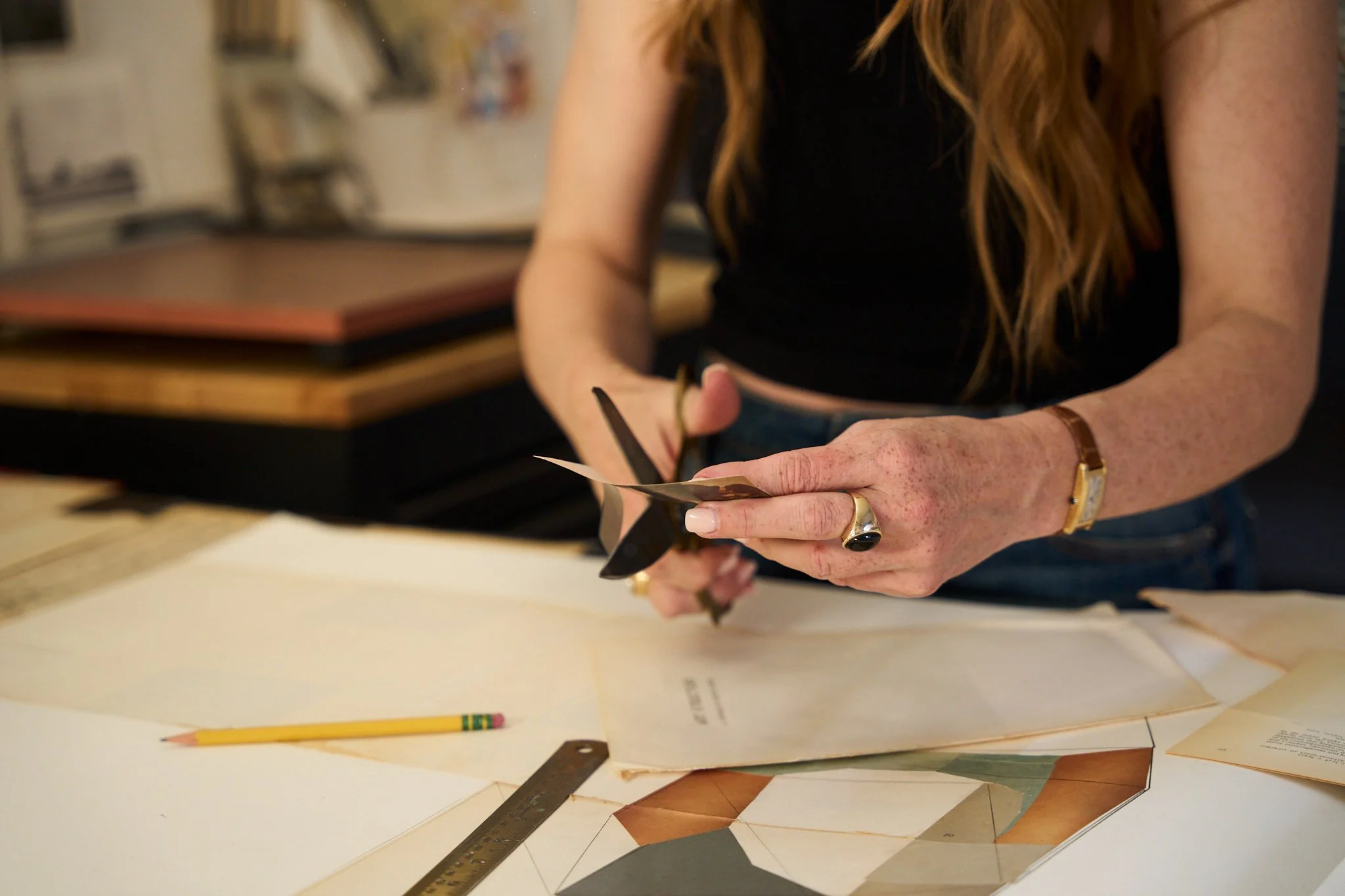 A person with long red hair, wearing a black sleeveless top, is working on an art project involving cut pieces of paper and a ruler on a table.