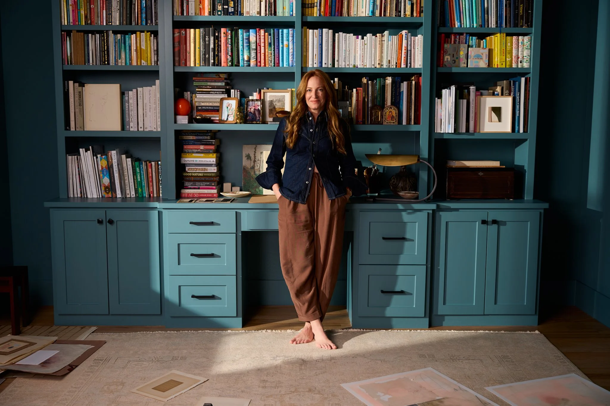Woman standing barefoot in front of a teal bookshelf filled with books and decorative items in a living room.
