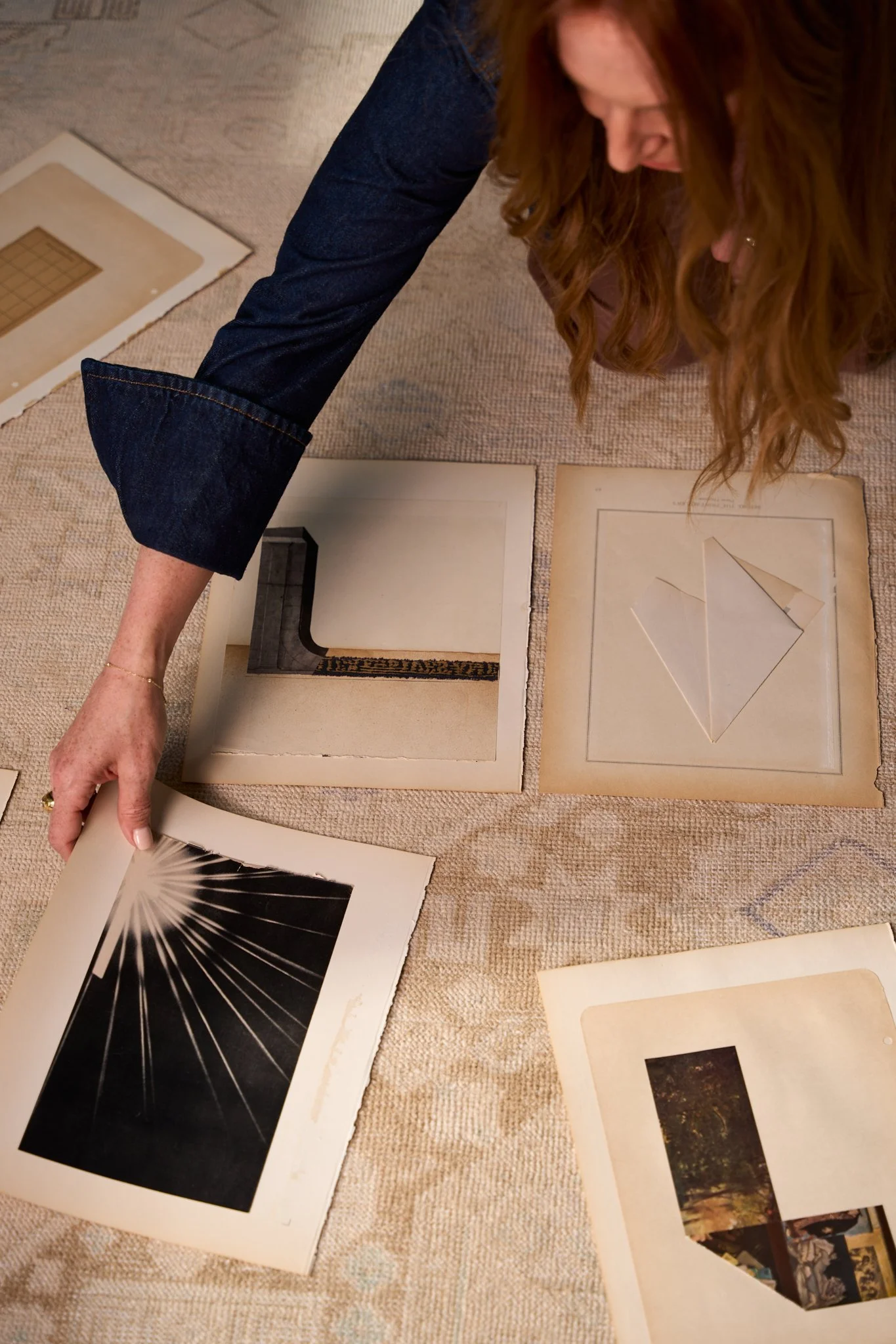 Person examining photographs and artwork laid out on a beige carpeted floor, including images of natural landscapes, abstract designs, and geometric patterns.