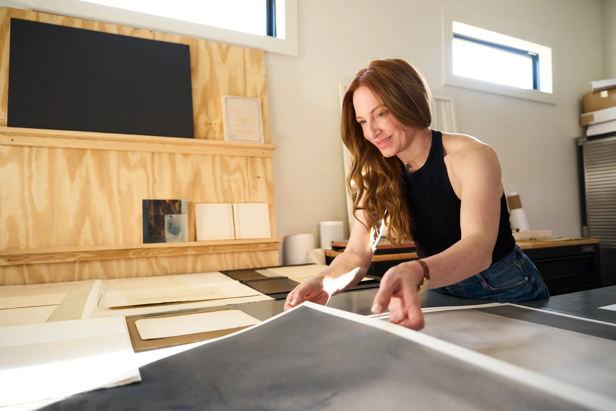 A woman with long red hair smiling while working on a project at a worktable in a well-lit room with wooden walls and large windows.