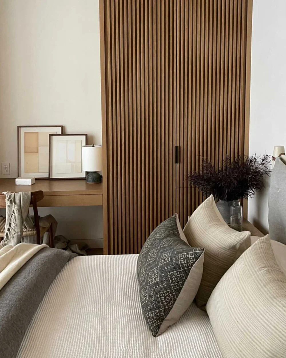Close-up of a bed with beige and dark patterned pillows, next to a nightstand with a vase and flowers, against a wooden slatted closet door.