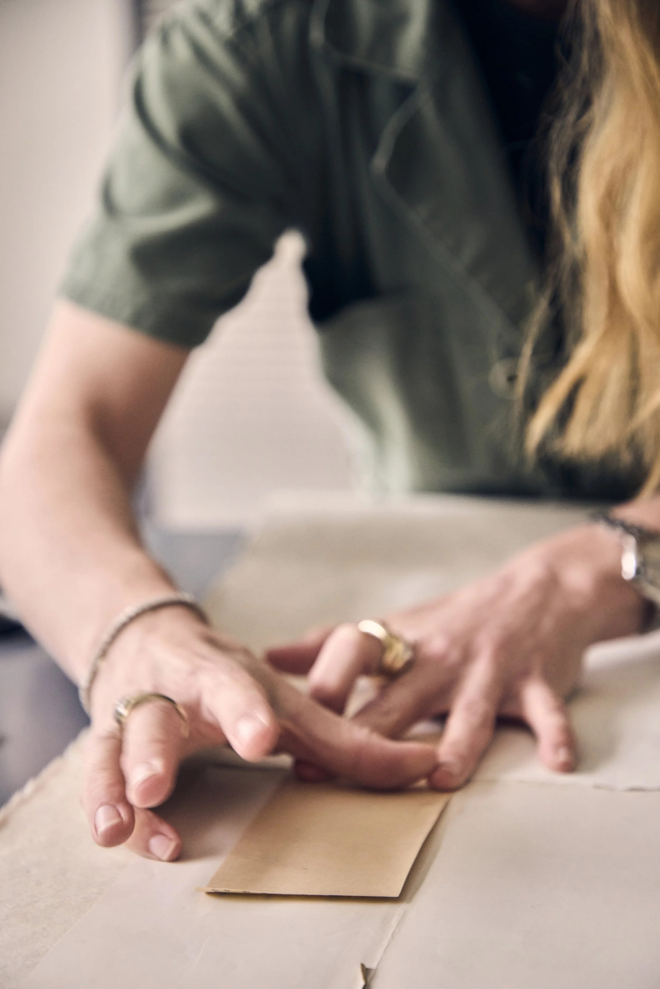 Close-up of a person's hands with rings, resting on a piece of paper on a table. The person is wearing a dark green shirt and has long, wavy blonde hair.