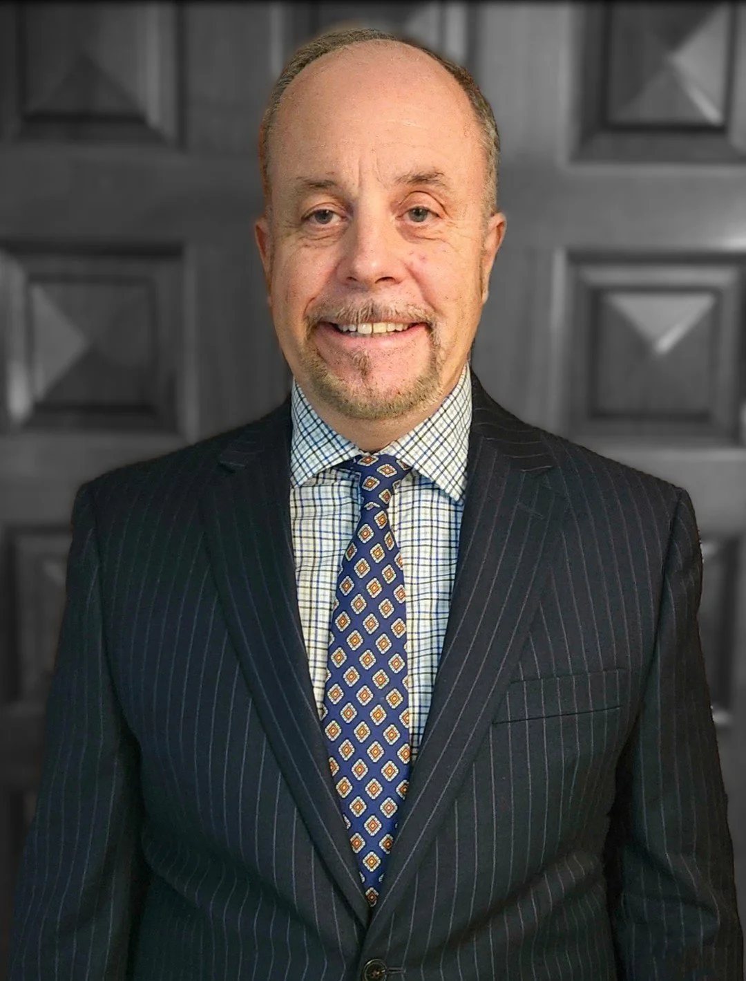 A man in a dark pinstripe suit, dress shirt, and patterned tie, smiling, in front of a gray wooden background.