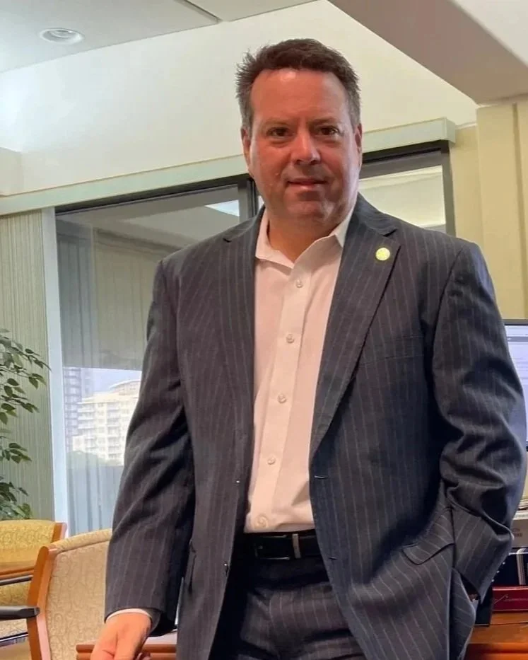 A man in a dark pinstripe suit and white dress shirt standing indoors near a window with buildings in the background.