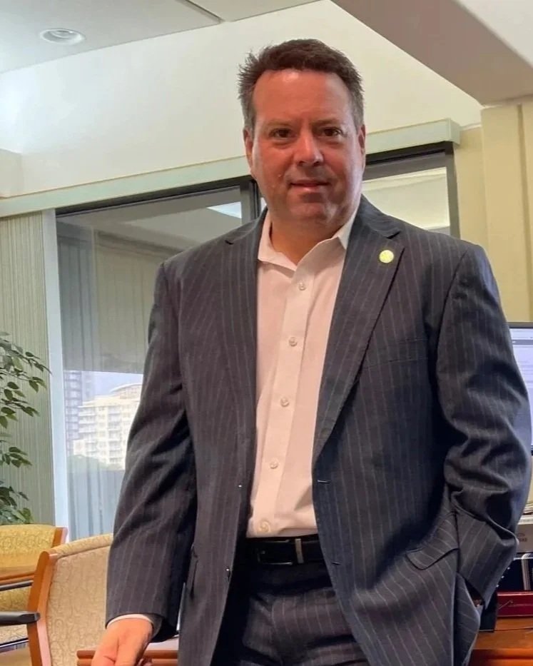 A man in a dark pinstripe suit stands in an office, with a white dress shirt underneath, and looks at the camera. There is a plant and a window with city buildings in the background.