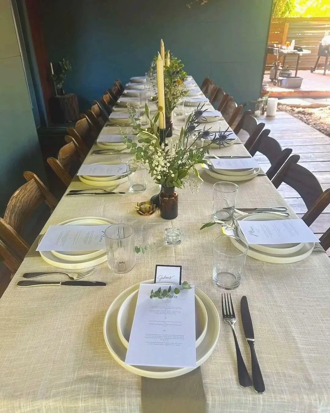Long dining table set for a formal meal with plates, silverware, glasses, and printed menus, decorated with flowers and candles in a bright outdoor or semi-outdoor space.