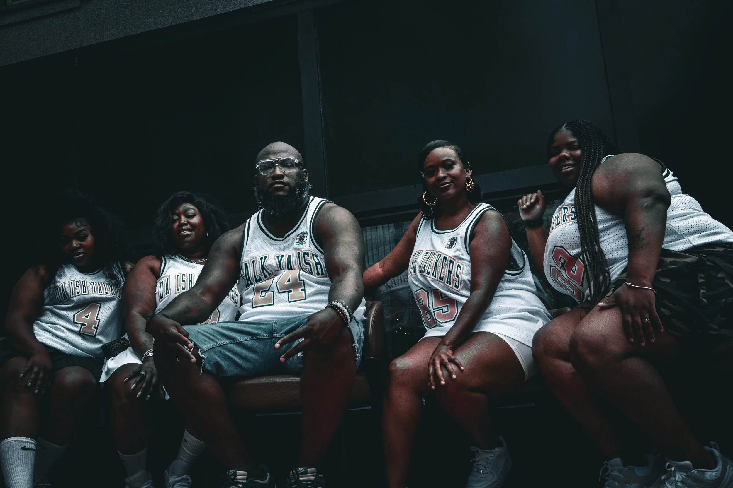 A group of five individuals in basketball jerseys sitting on a couch outdoors, posing for a photo.
