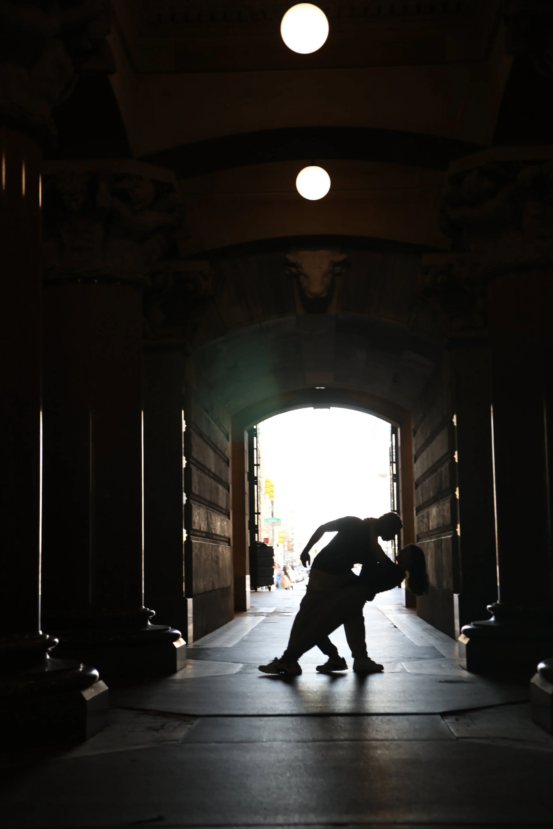 Silhouette of a man and a girl sharing a kiss in a doorway, backlit by outdoor daylight in an architectural archway with decorative sculptures on the ceiling.