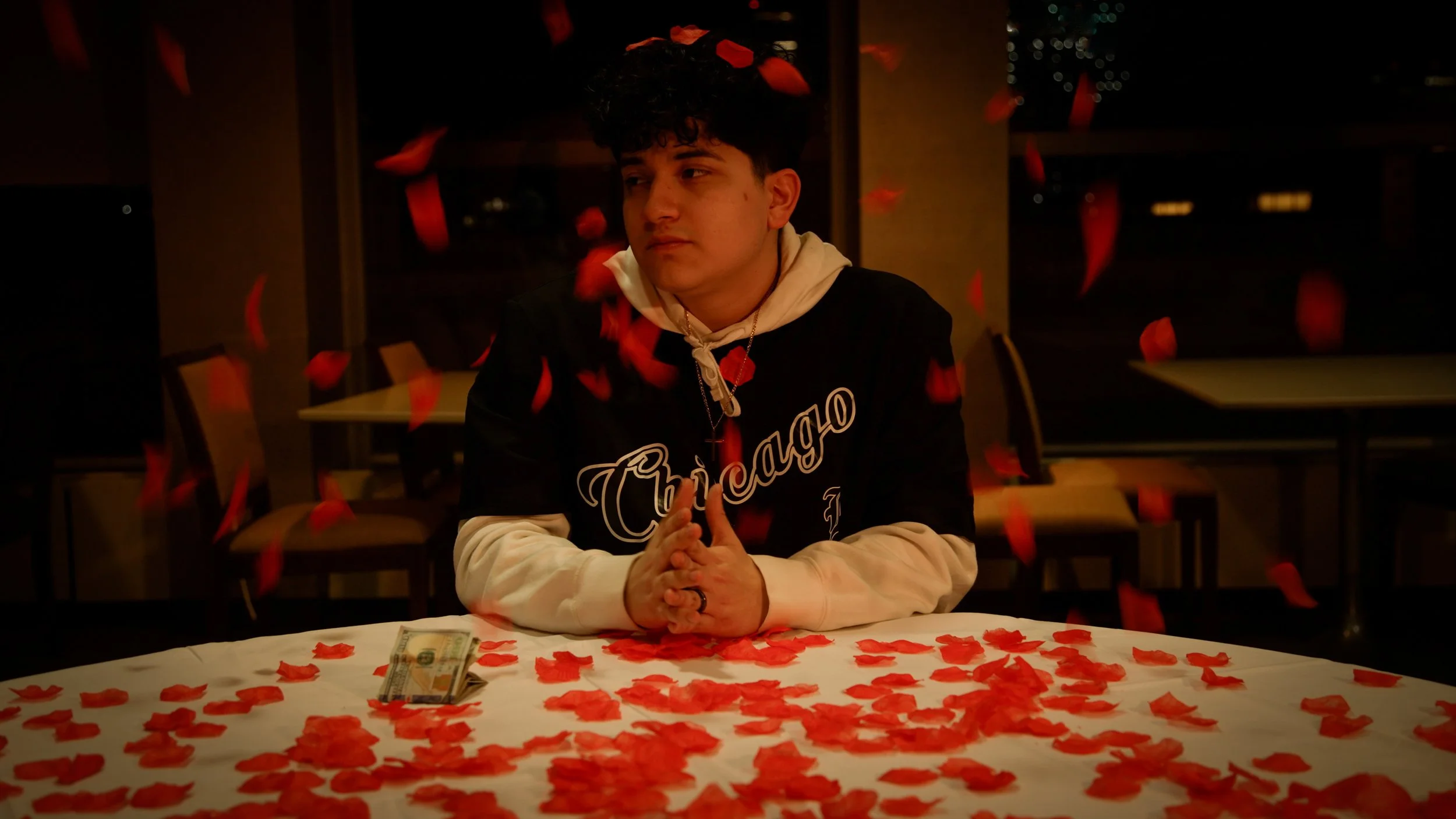 A young man with curly hair sitting at a table covered with red rose petals, with a stack of hundred-dollar bills in front of him. Blue petals are falling around him in a dimly lit room with a window in the background.