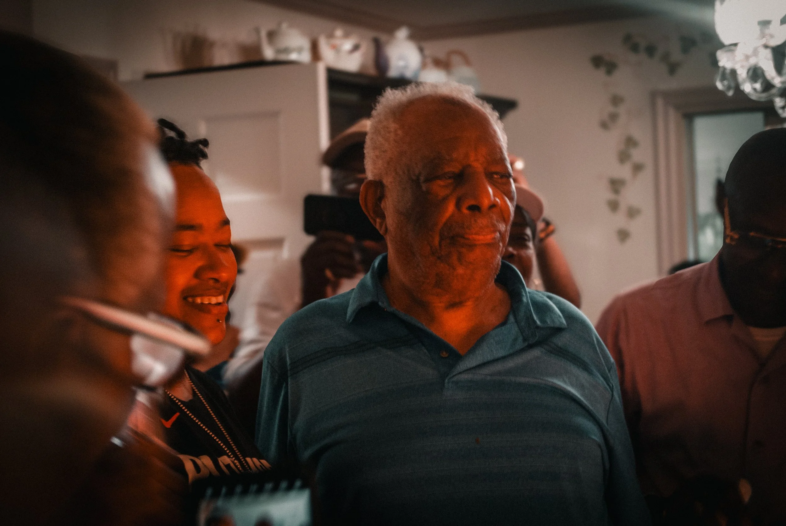 An elderly man with gray hair and a blue striped polo shirt stands among a group of smiling people at an indoor gathering.