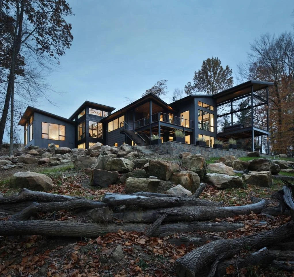 Modern black house with large windows, situated on a rocky, wooded hillside during dusk.