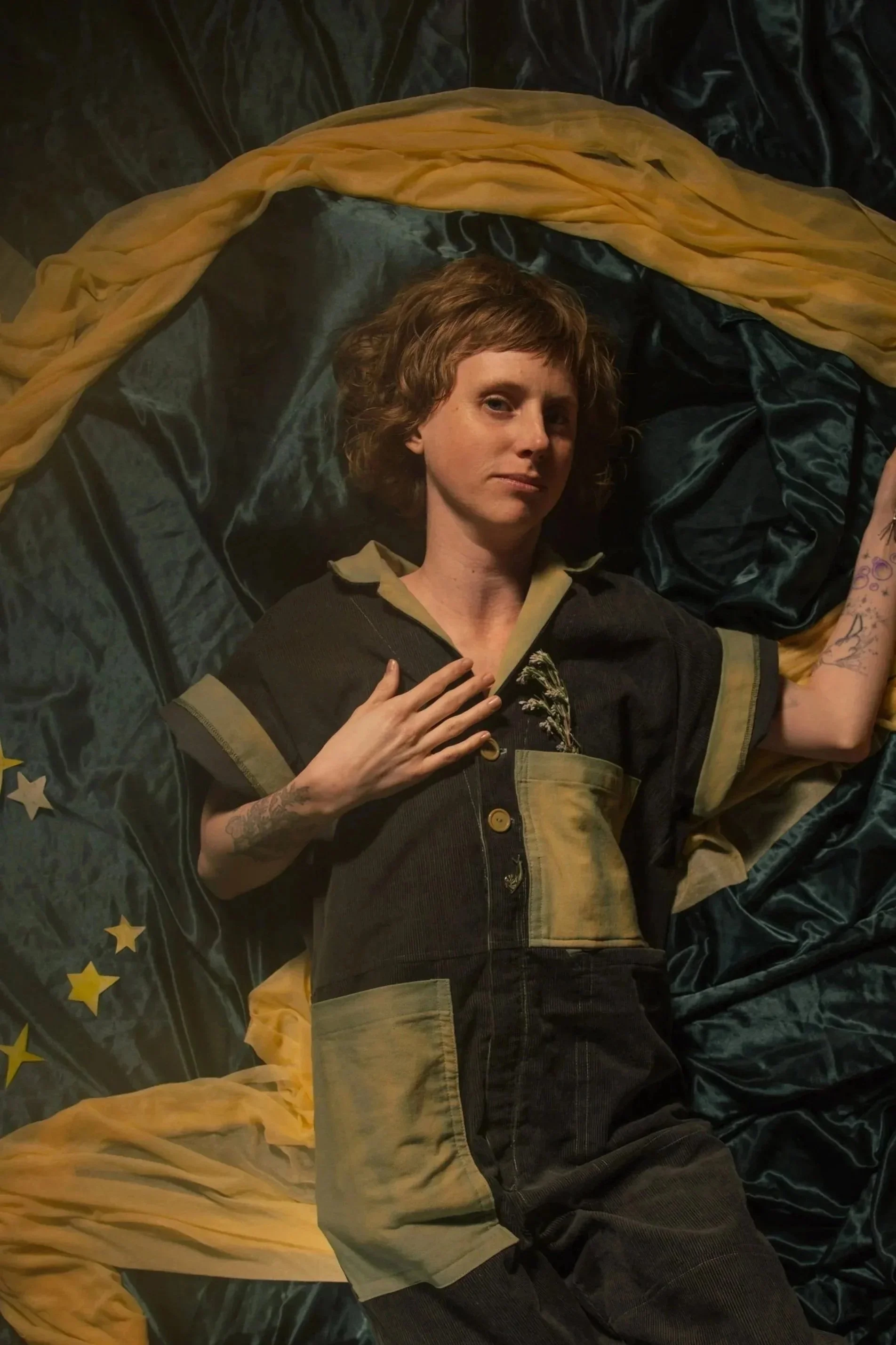 Person reclining on dark fabric beneath draped mustard-yellow cloth, wearing a dark outfit with olive-green accents and visible tattoos.