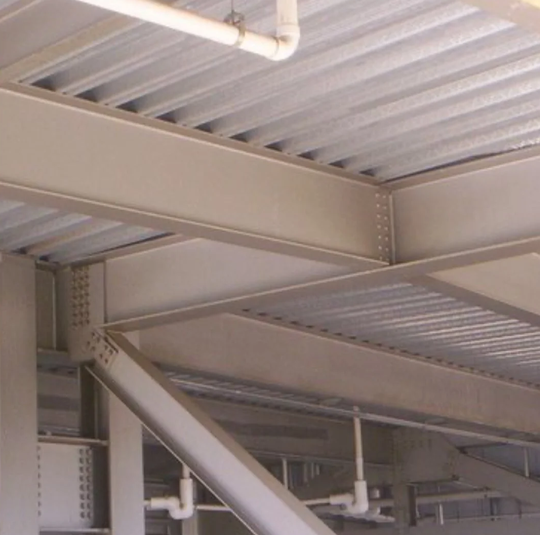 Interior view of a metal structure with beams, pipes, and a corrugated metal roof.