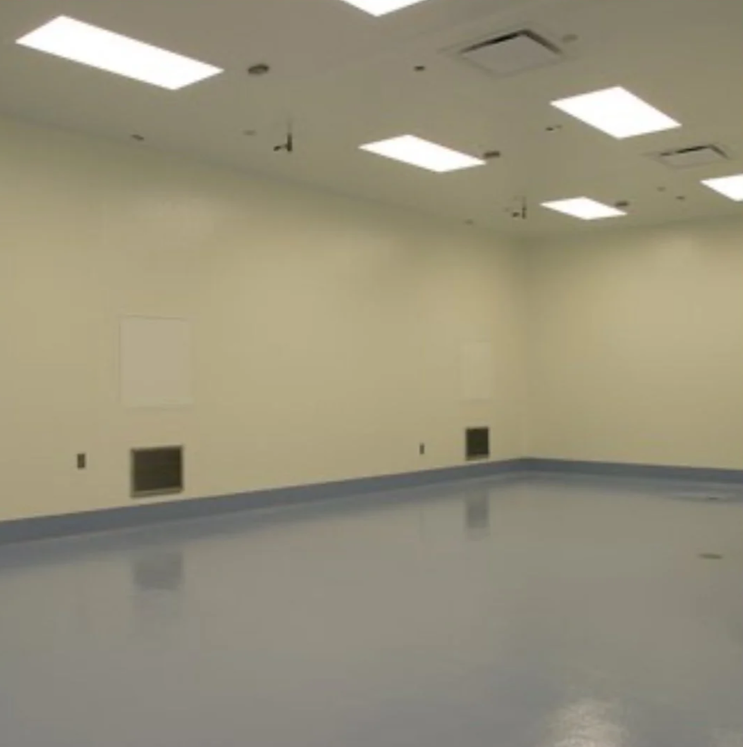 Empty room with beige walls, gray baseboards, and a shiny gray floor. The ceiling has multiple rectangular fluorescent lights and an air vent.