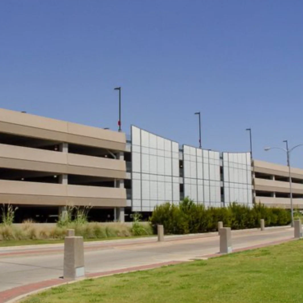A multi-story parking garage with beige walls, concrete supports, and an exterior glass facade, with bushes and grass in the foreground and a clear blue sky overhead.