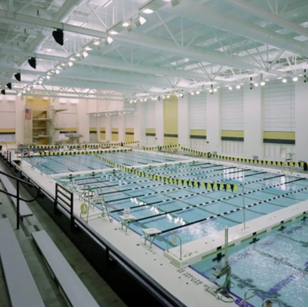 Indoor swimming pool with multiple lanes marked by black and yellow lane dividers, surrounded by white walls and large windows with shades, and lounge chairs along the poolside.