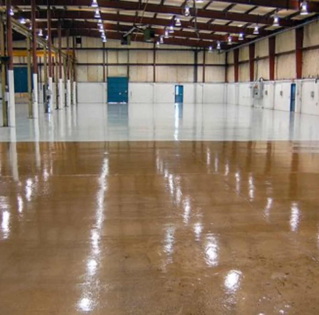 Interior of an empty warehouse with polished wooden and white concrete floors, high ceiling with exposed beams and hanging lights, and blue doors along the back wall.