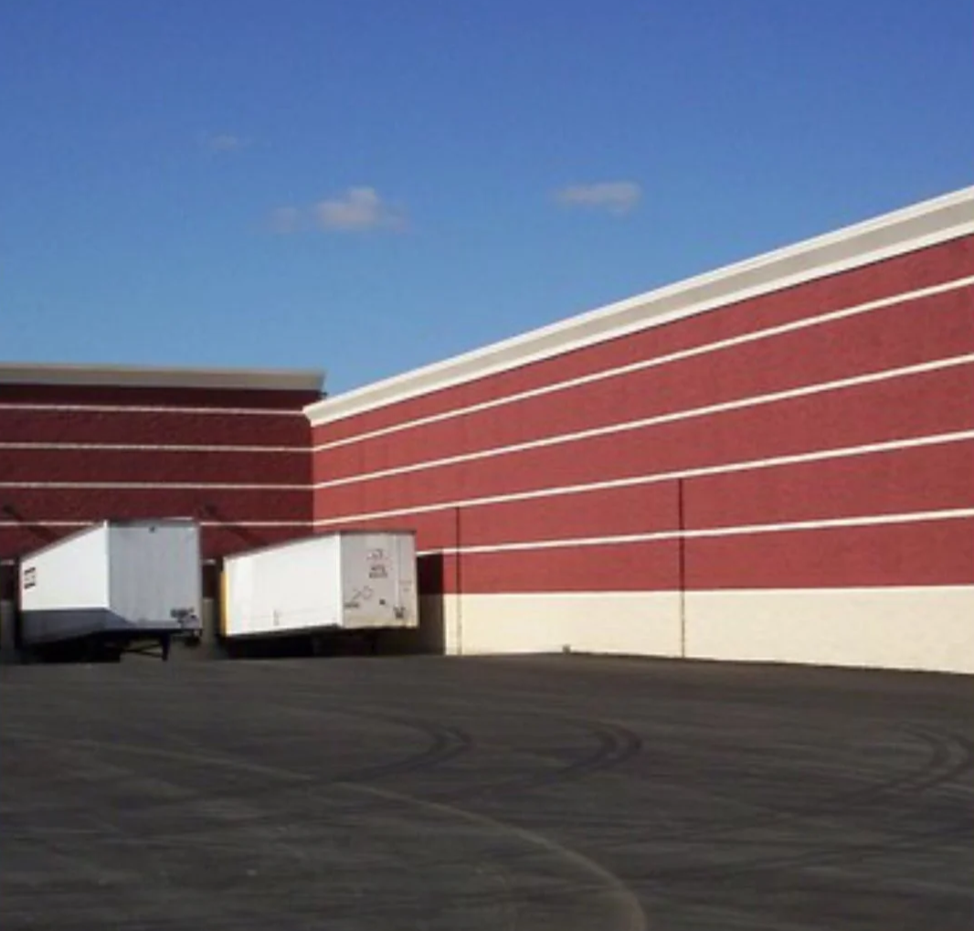 Red brick building with white horizontal lines, three white loading dock doors, and a paved parking lot with tire marks under a blue sky with a few clouds.