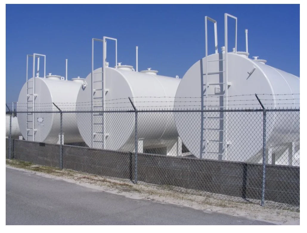 Three large white storage tanks behind a chain-link fence with barbed wire, set against a bright blue sky.