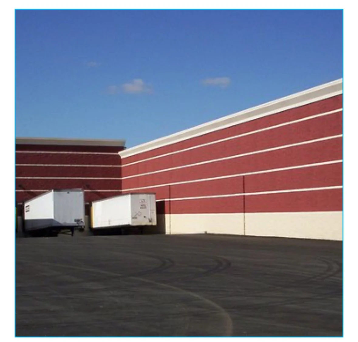 Red brick building with white horizontal lines, three white loading dock doors, and a paved parking lot with tire marks under a blue sky with a few clouds.