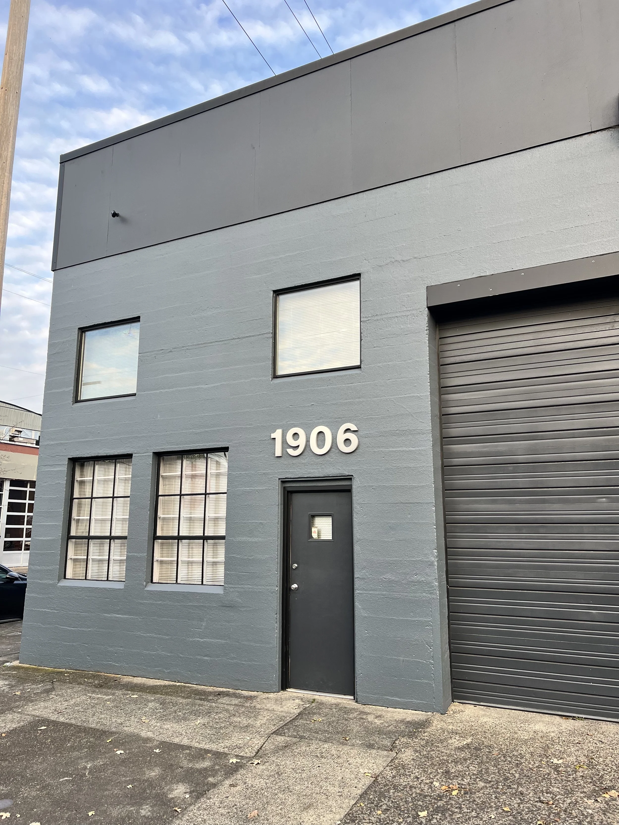Gray commercial building with the number 1906 on the wall, two windows with blinds, a solid black door, and a large gray roll-up door, with a concrete sidewalk in front.