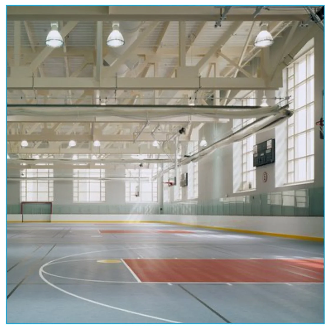 Empty indoor ice hockey rink with goal net, ice surface, and large windows letting in natural light.