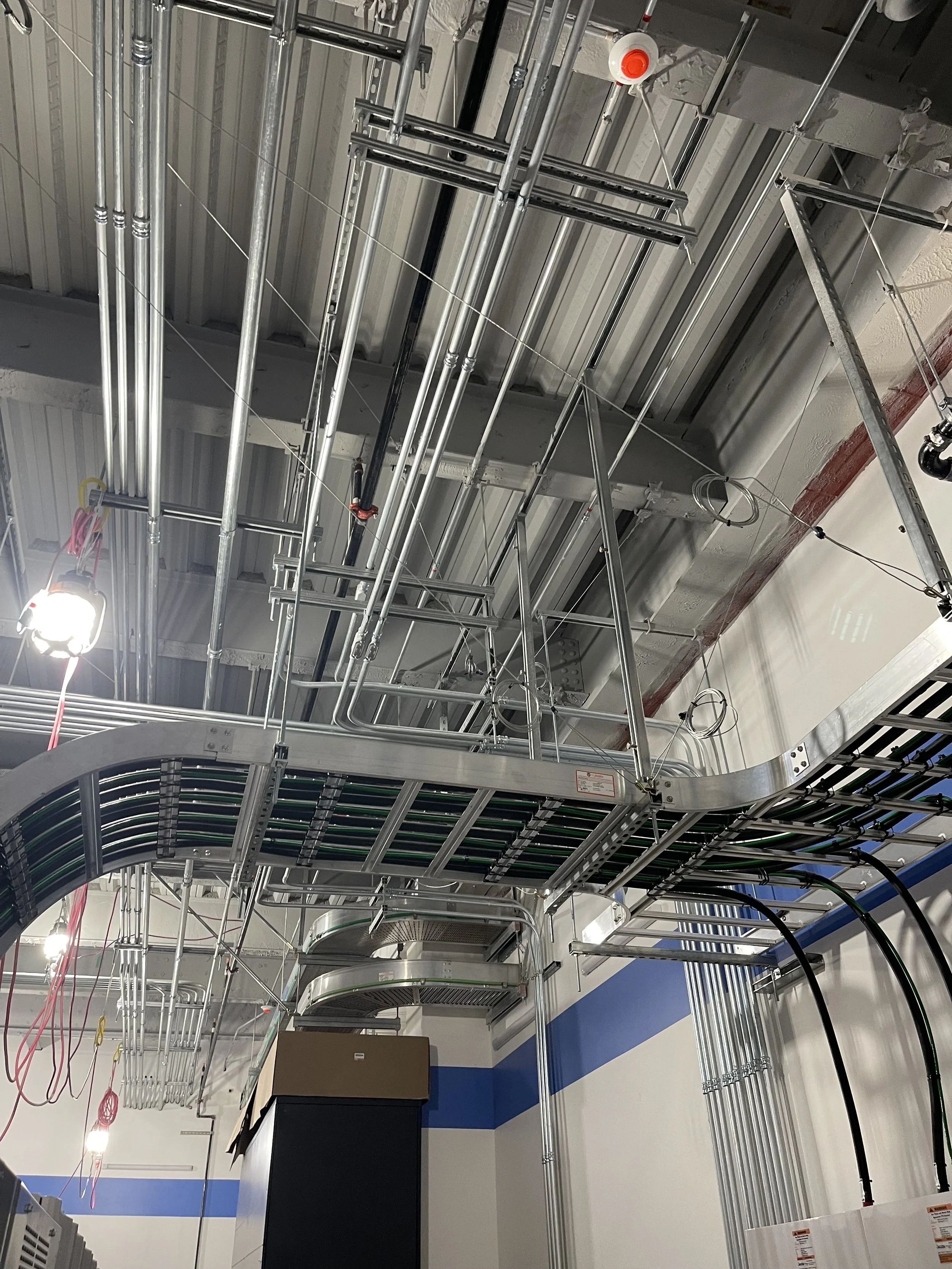 View of a ceiling under construction with exposed metal pipes, conduits, and framing, along with temporary lighting and electrical wiring in a building interior.