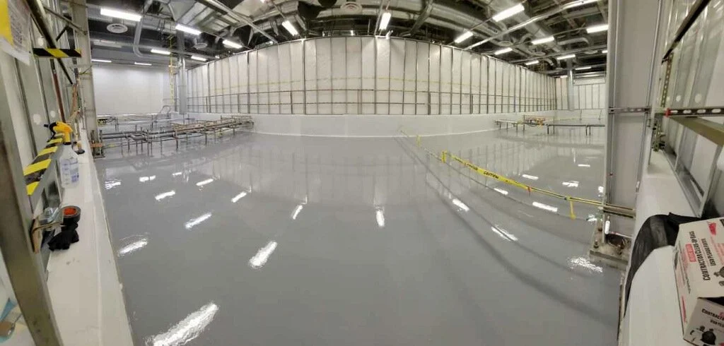 Empty indoor industrial space with a shiny, polished concrete floor, metal railings, and a high ceiling with structural supports.