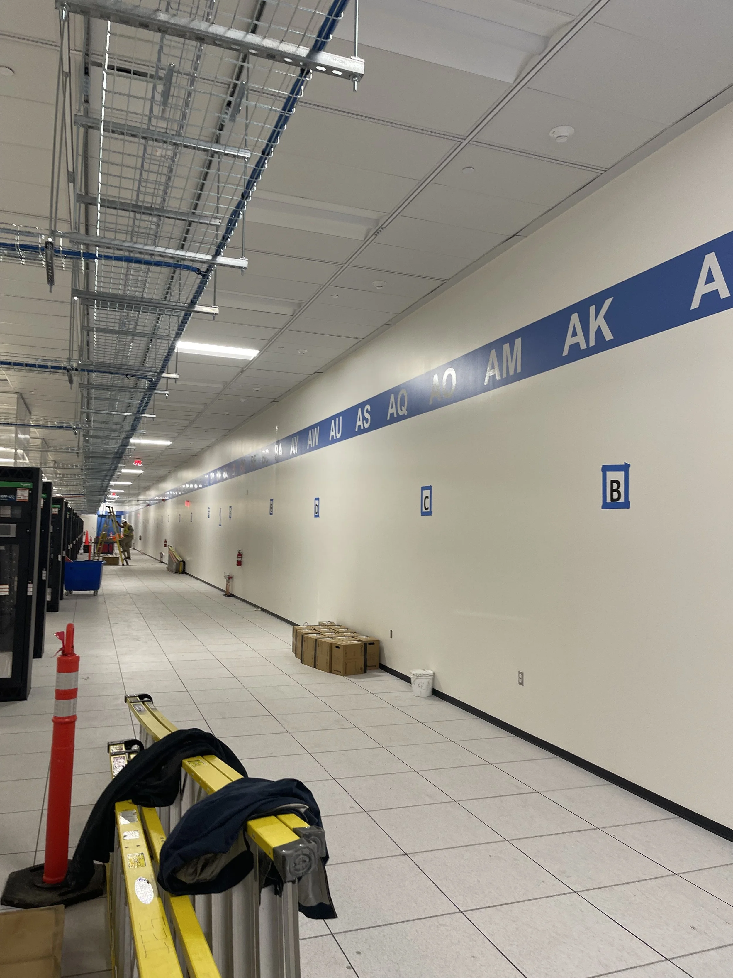 An indoor corridor under construction with a blue and white sign on the wall indicating airline gates such as AA, AQ, AU, AS, and AK. Worker in the background, boxes on the floor, construction materials on ceiling, and yellow safety barriers in the f