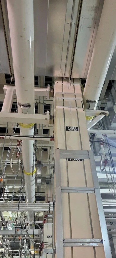 Industrial ceiling with large metal pipes, support beams, and a wooden ladder marked 'AB-56'.