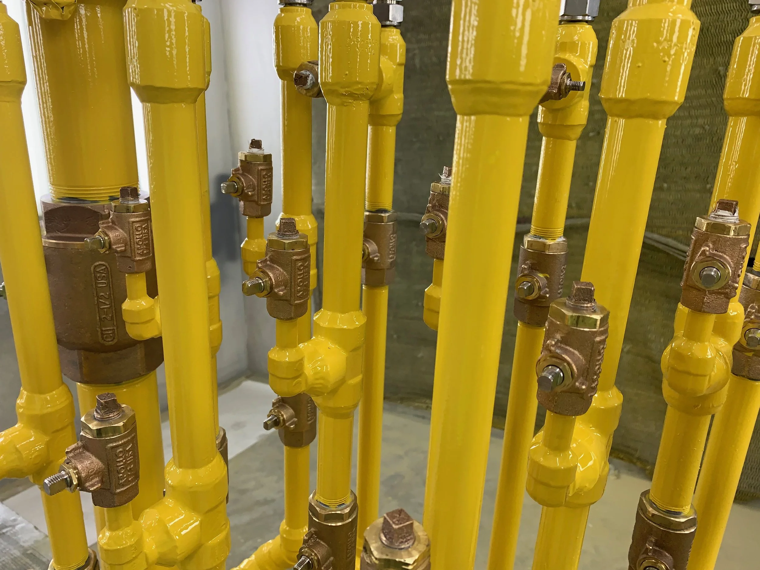 A series of yellow painted pipes connected with bronze fittings and valves, installed vertically on a wall.