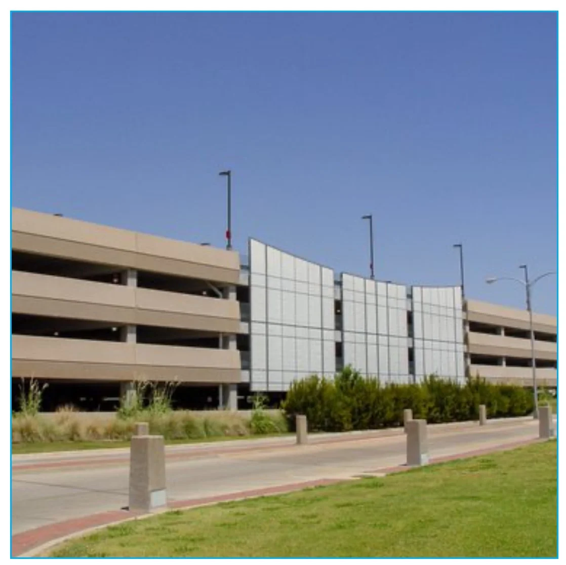 A multi-story parking garage with beige walls, concrete supports, and an exterior glass facade, with bushes and grass in the foreground and a clear blue sky overhead.