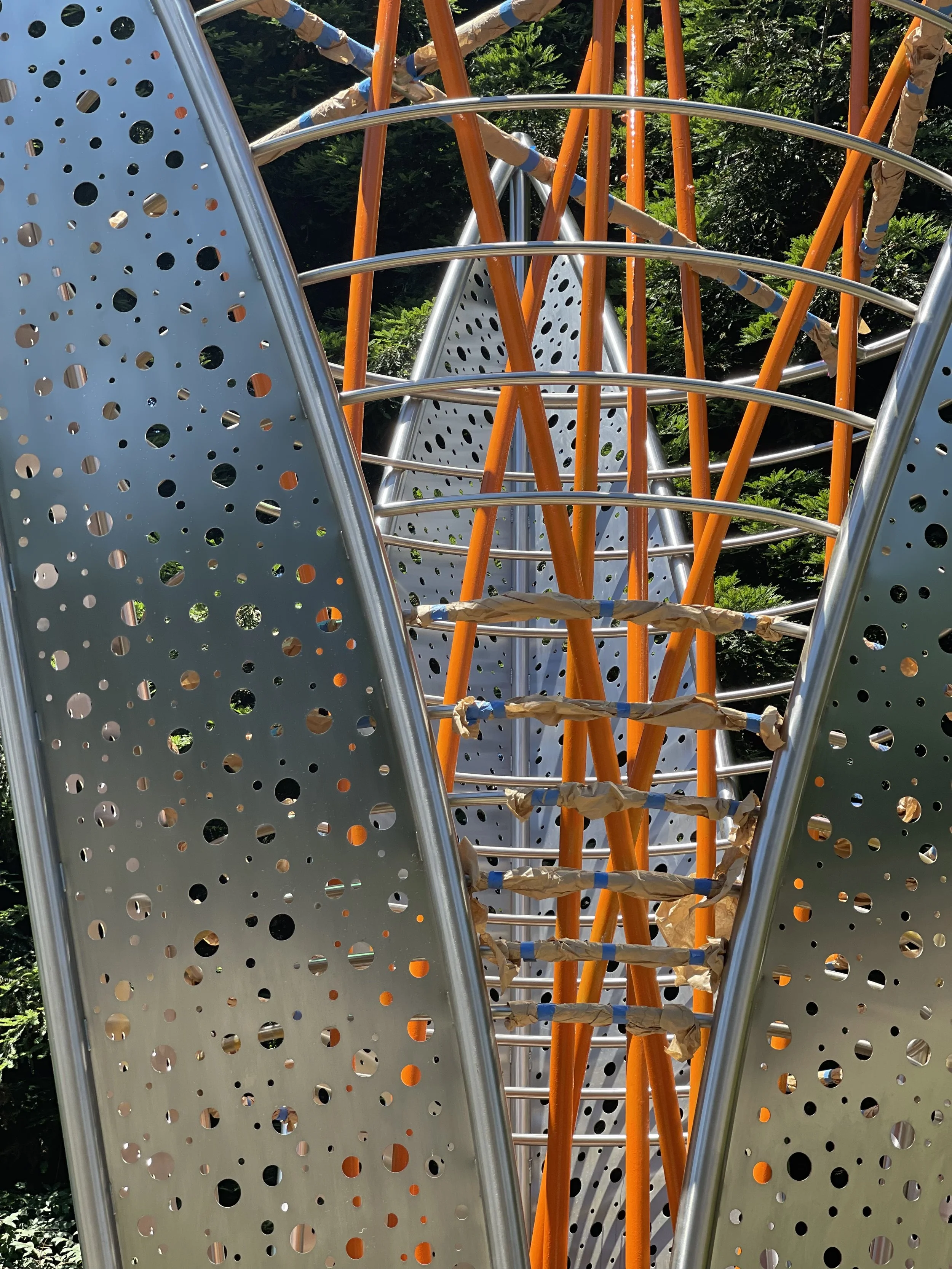 Close-up view of a modern playground climbing structure with metal and orange painted bars, surrounded by greenery.