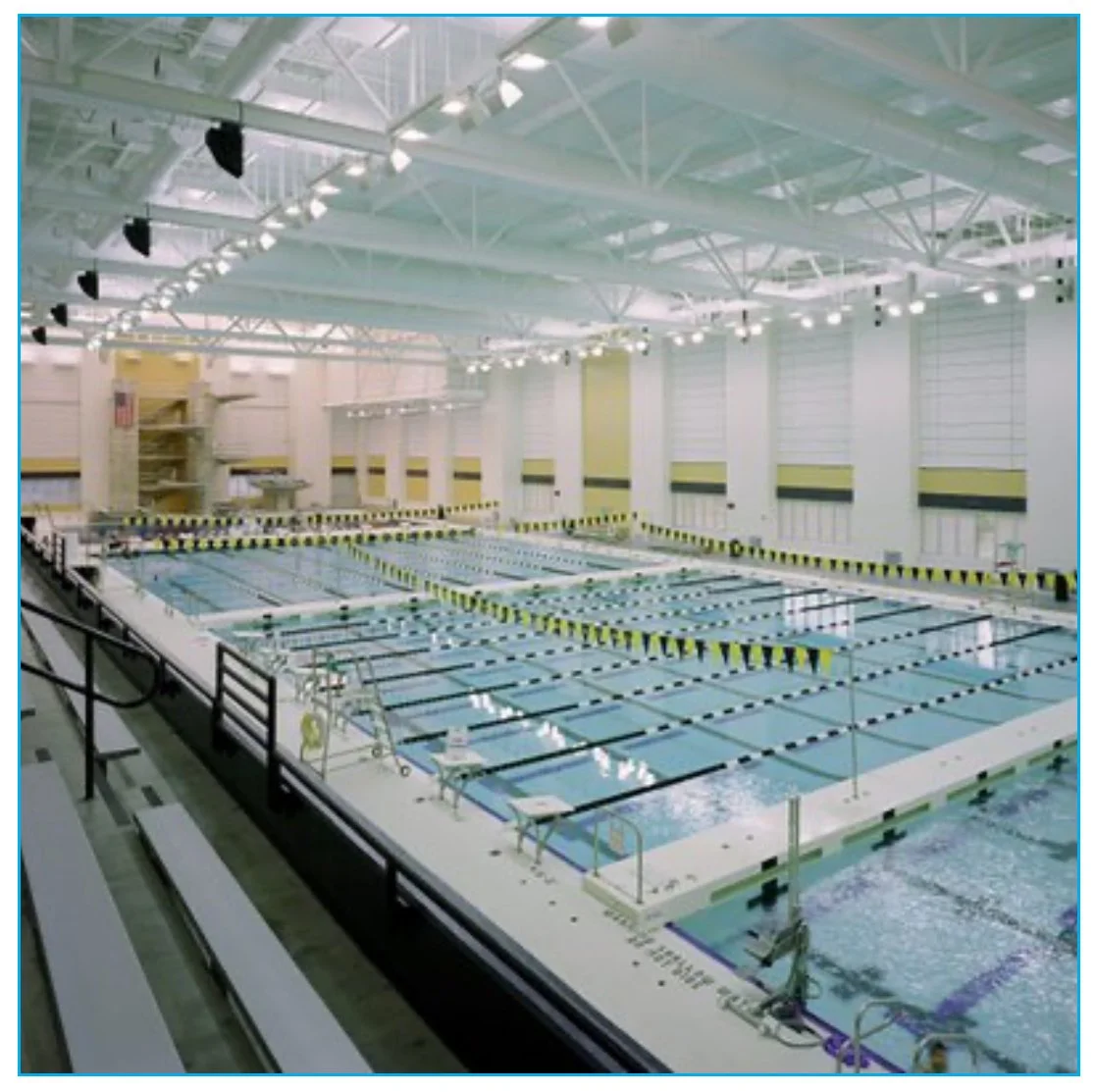 Indoor swimming pool with multiple lanes marked by black and yellow lane dividers, surrounded by white walls and large windows with shades, and lounge chairs along the poolside.