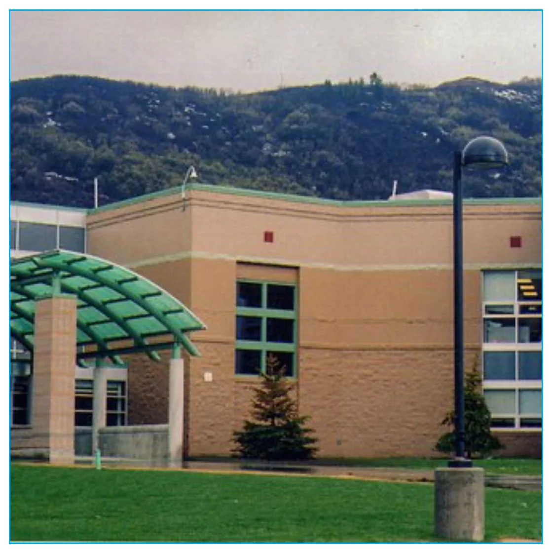 Image of a school or office building with a mountain in the background, trees, a grass lawn, a streetlamp, and a green glass awning over the entrance.
