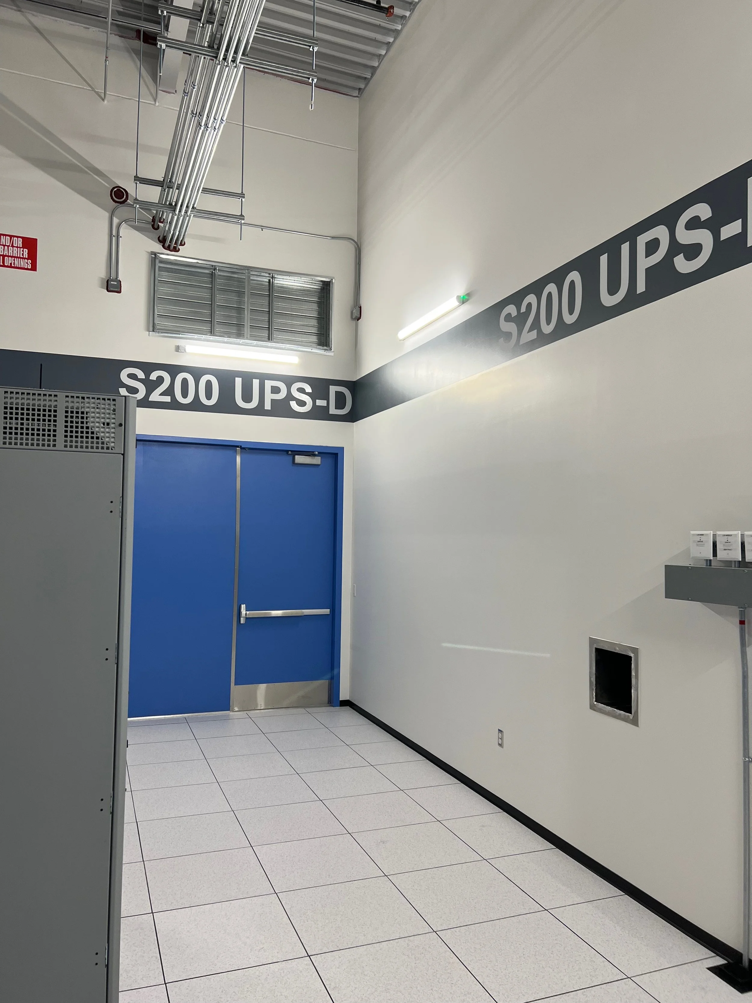 An industrial room with white walls, gray tiled flooring, a blue door, and signage reading 'S200 UPS-D' on the wall. There are electrical conduits and piping on the ceiling, a vent, and some small boxes or labels on a gray panel on the right.