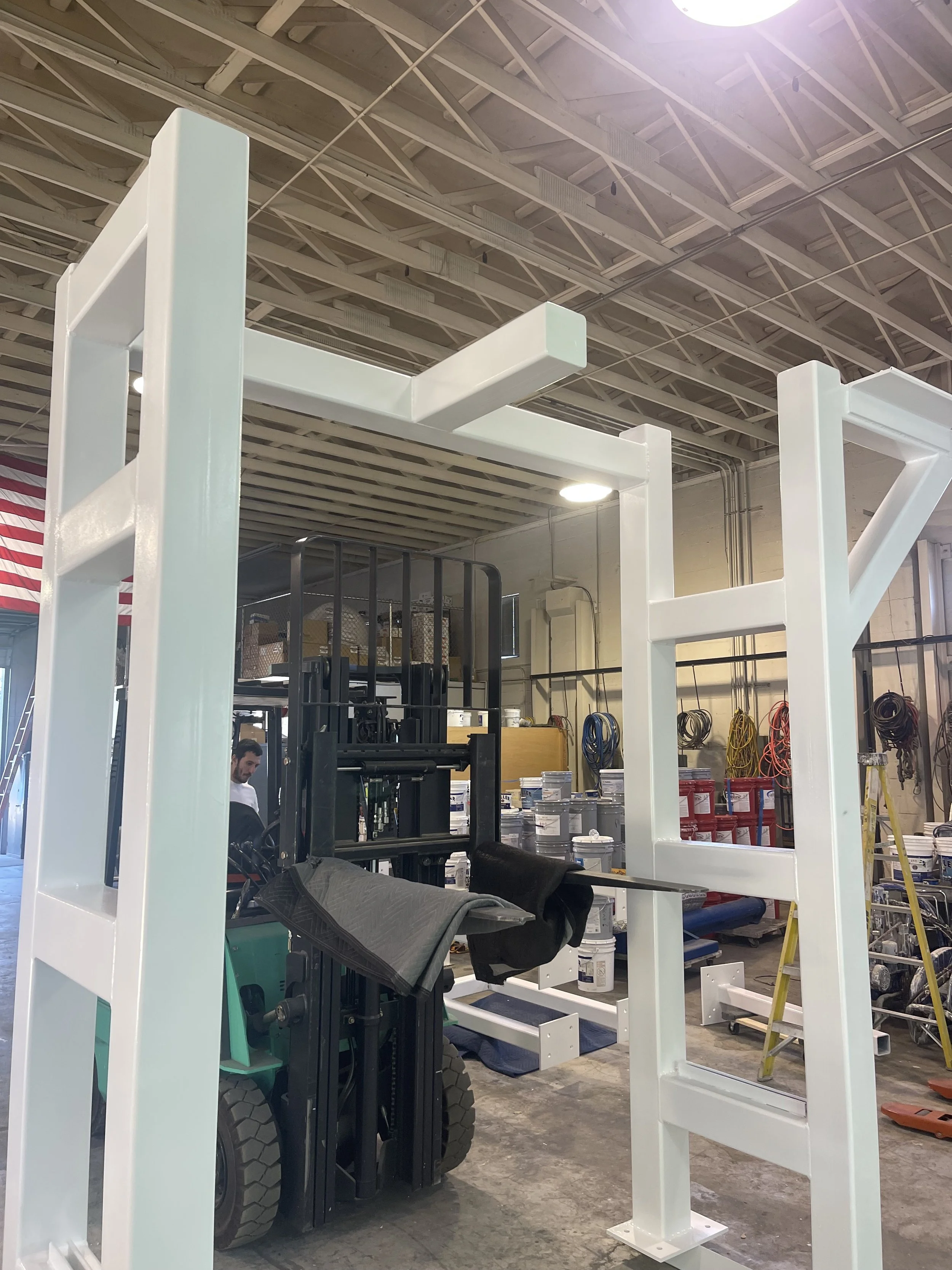 Industrial warehouse with a large white metal frame structure, a black forklift, and shelves filled with buckets and equipment. Worker in the background.