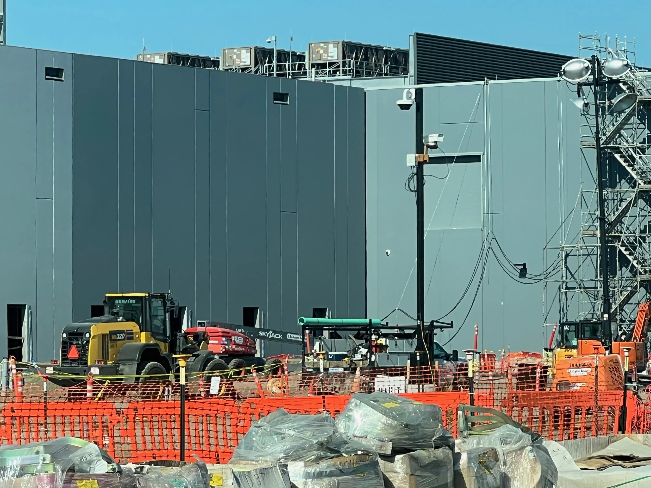 Construction site with orange safety fencing, green pipes, a yellow and black loader, and a gray building with rooftop equipment.